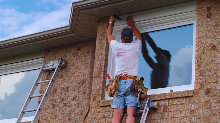 A homeowner on a ladder with a tool belt trying to install a new window in NJ.